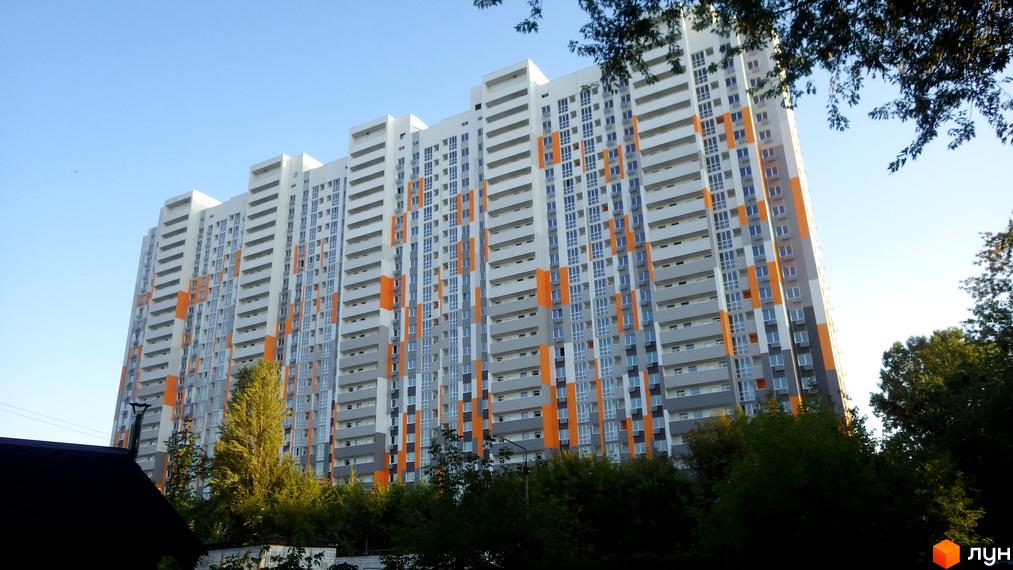 Зовнішній вигляд ЖК Пектораль: висока багатоповерхова будівля з білими, сірими та помаранчевими елементами фасаду. Навколо будівлі зелені дерева та блакитне небо.