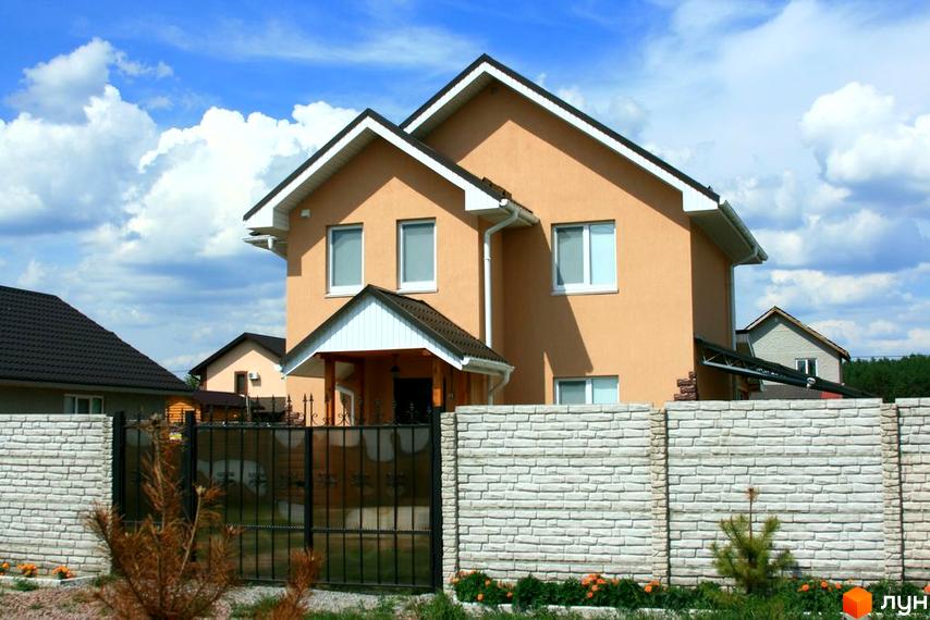 Котеджне містечко КМ Лісна Пісня. Двоповерховий будинок світло-бежевого кольору з білими вікнами та дахом. Огороджений цегляним парканом та кованими воротами.