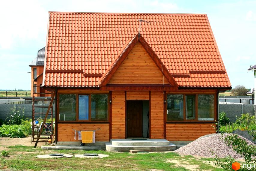 Зображення будинку в Котеджному містечку Лісна Пісня з червоним дахом. Навколо будинку зелений газон та невелика ділянка з гравієм.