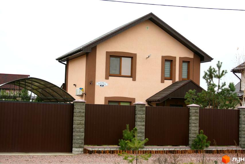 Зображення двоповерхового будинку з коричневим дахом та парканом. Вигляд фасаду та прибудинкової території Котеджного містечка Лісна Пісня.