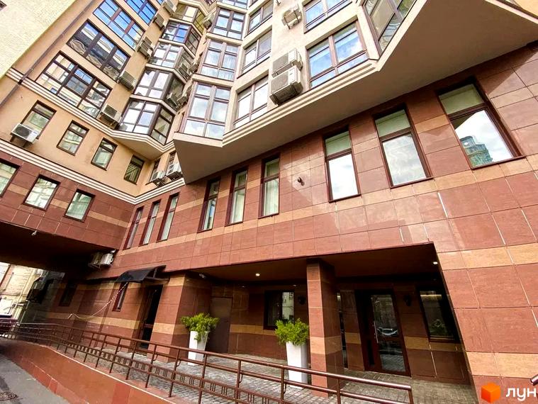 Багатоповерховий будинок Житлового комплексу Botanic Paradise з фасадом, облицьованим коричневими та бежевими плитками, та численними вікнами. Перед входом облаштований пандус з поручнями та зелені насадження у вазонах.