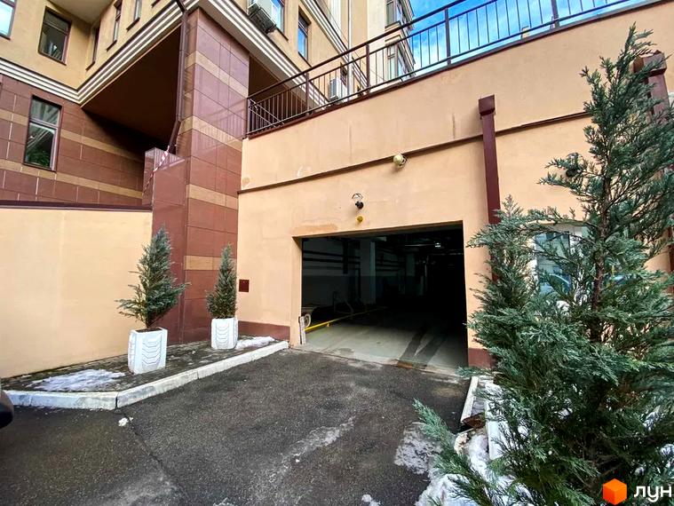 Вхід до підземного паркінгу Житлового комплексу Botanic Paradise. Будівля має бежевий фасад з коричневими елементами, поруч ростуть хвойні дерева.