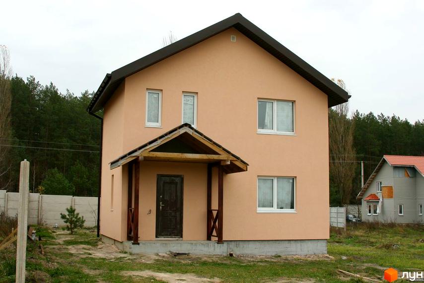 Двоповерховий будинок з мансардою та ґанком. Навколо будинку зелена трава та дерева, неподалік розташована інша будівля.