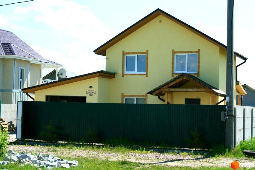 Фото двоповерхового будинку з жовтими стінами та коричневим дахом в Котеджному містечку Лісна Пісня. На передньому плані зелений паркан та газон.