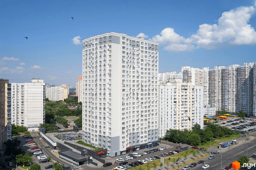 Висотна будівля житлового комплексу з великою кількістю вікон. На прибудинковій території є парковка для автомобілів та зелені насадження.