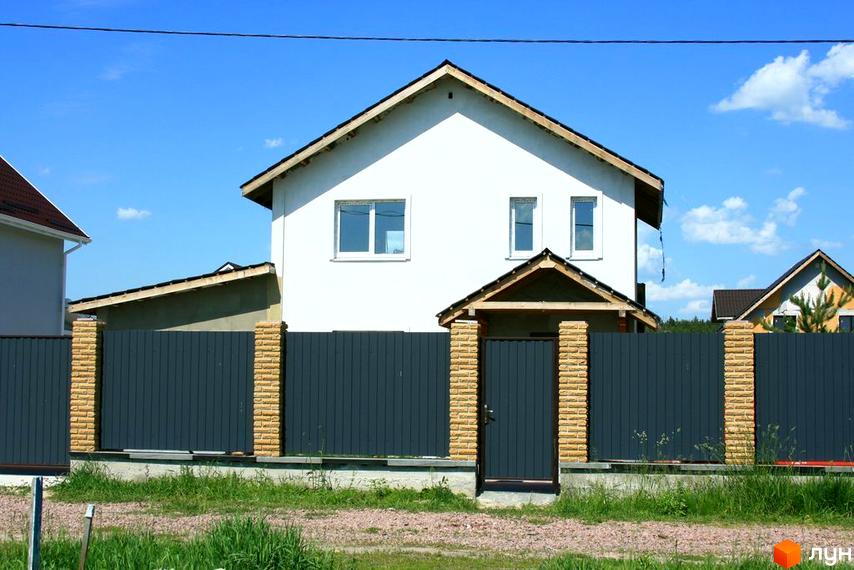 Двоповерховий будинок з білими стінами та темним дахом. Обгороджений сірим парканом з цегляними стовпчиками та хвірткою.