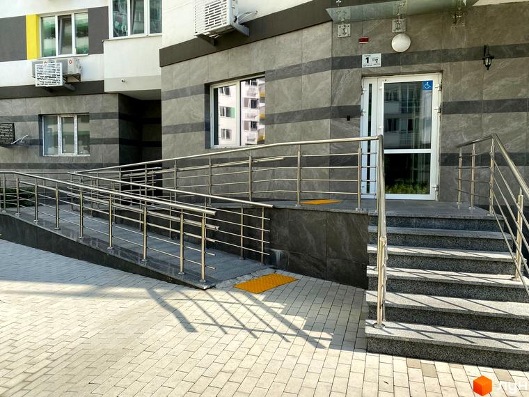 Зовнішній вигляд входу до житлового комплексу Харківський з пандусом та сходами. Прибудинкова територія викладена плиткою.