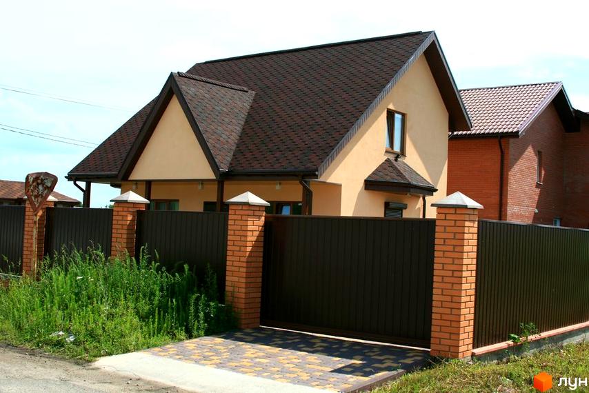Вигляд будинку в Котеджному містечку Лісна Пісня з коричневим дахом та огорожею. Подвір'я з тротуарною плиткою та зеленою травою.