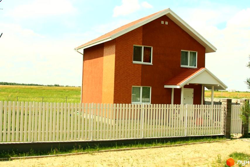 Фото двоповерхового будинку з червоною цеглою та білим парканом у Котеджному містечку Лісна Пісня. Навколо будинку зелений газон та поле.