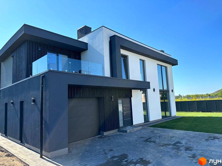 Сучасний двоповерховий будинок з великими вікнами та скляним балконом, розташований на доглянутій прибудинковій території з газоном та бруківкою.