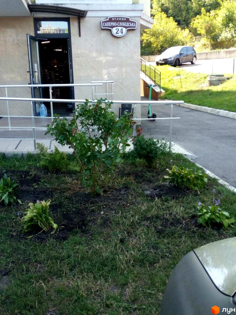 Вхід до житлового комплексу на вул. Саперно-Слобідська, 24, з рослинністю на прибудинковій території.