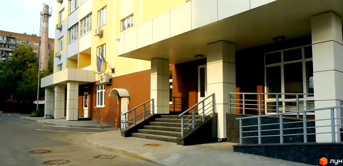 Клубний будинок Gelios має жовто-коричневий фасад з білими колонами та навісом над входом. Біля будинку розташовані сходи з перилами та прибудинкова територія з асфальтованим покриттям.