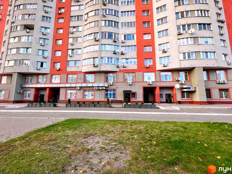 Фасад Житлового комплексу Воскресенський з сірими та червоними елементами, великими вікнами. Прибудинкова територія має доріжки та зелений газон.