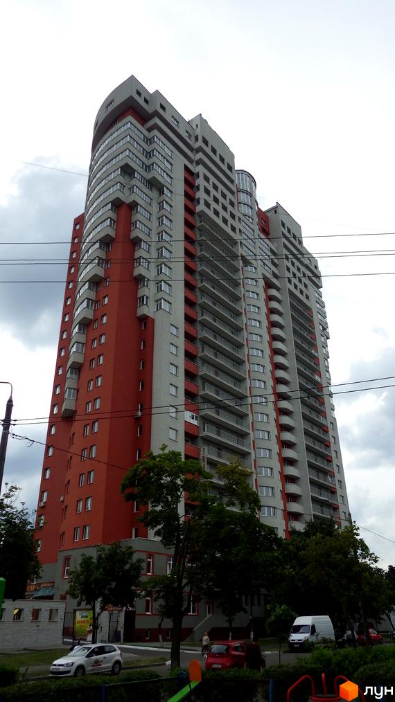 Багатоповерховий житловий будинок Житловий комплекс Воскресенський з фасадом червоного та світло-сірого кольорів, з численними балконами. Навколо будинку видно дерева та дорогу.