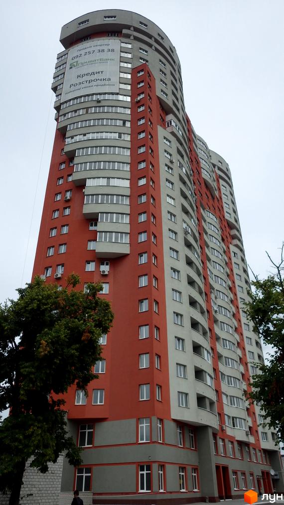 Багатоповерховий житловий комплекс Воскресенський з червоно-сірим фасадом та округлими балконами. Навколо будинку видно зелені дерева.