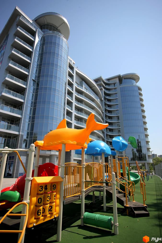 Багатоповерхові будівлі Житлового комплексу Рів'єра Ріверсайд зі скляними фасадами та округлими балконами. На передньому плані яскравий дитячий майданчик з гірками та ігровими елементами.