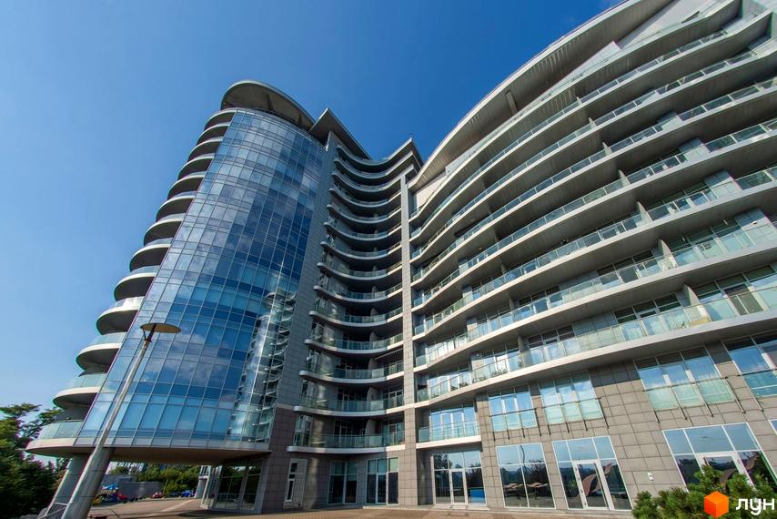 Багатоповерховий житловий комплекс Рів'єра Ріверсайд з вигнутим скляним фасадом та балконами. Навколо будівлі видно тротуар та зелені насадження.