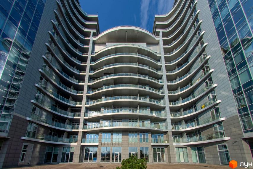 Фасад Житлового комплексу Рів'єра Ріверсайд з вигнутими лініями, скляними балконами та великими вікнами. На передньому плані видно вимощену територію.