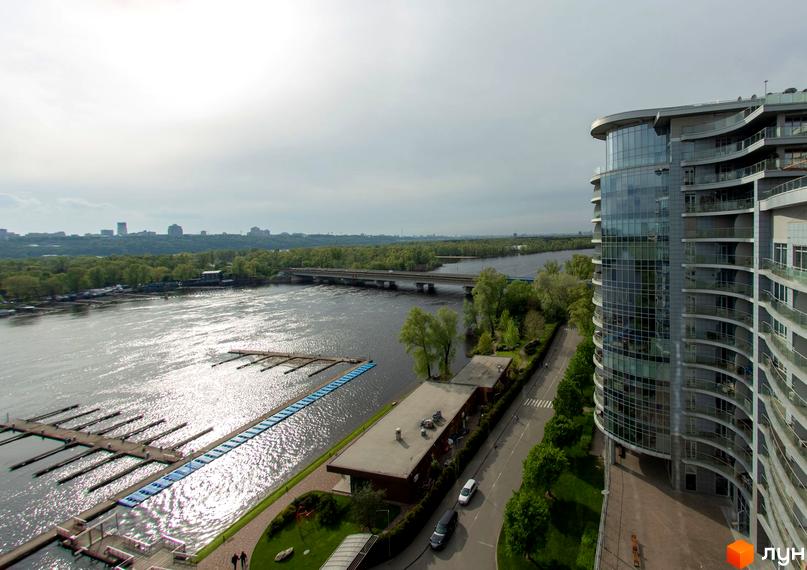 Житловий комплекс Рів'єра Ріверсайд з вигнутим скляним фасадом та балконами, розташований біля річки з причалами та зеленими насадженнями.