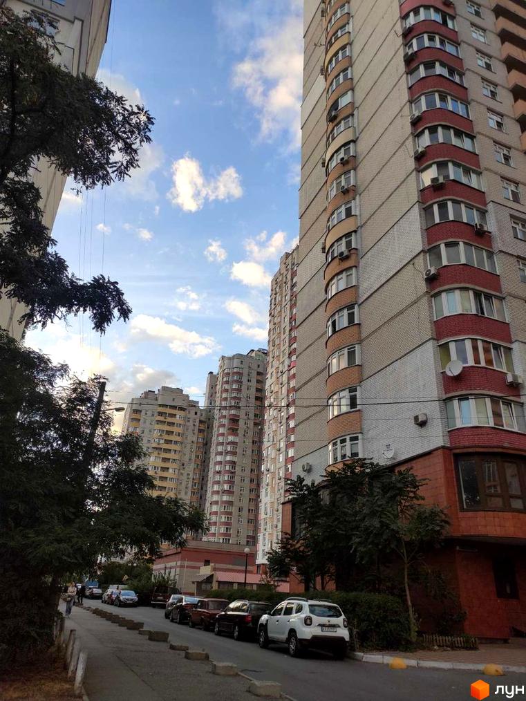 Багатоповерховий житловий будинок зі світлої цегли з червоними та помаранчевими балконами. Поруч вулиця з припаркованими автомобілями та зеленими насадженнями.