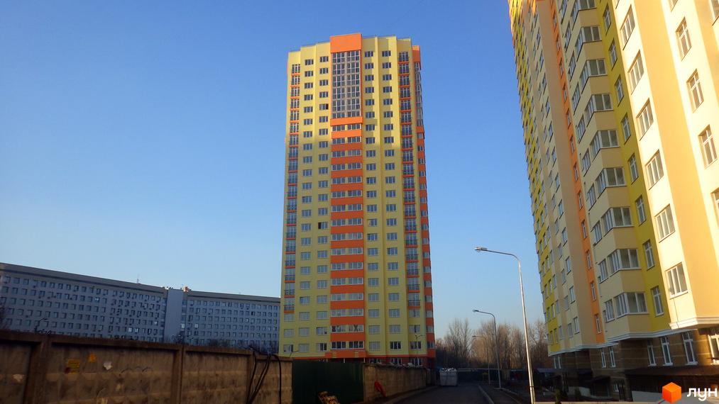 Житловий комплекс Яскравий: висотні будинки з жовто-помаранчевим фасадом та великою кількістю вікон. Поруч розташована сіра багатоповерхова будівля. На передньому плані - паркан та дорога.