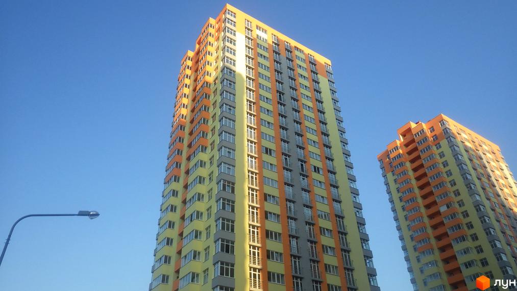 Житловий комплекс Яскравий - це сучасна новобудова з яскравим фасадом, що складається з висотних будинків. На зображенні видно два будинки з жовтими та помаранчевими елементами, що доповнюються сірими вставками. Перед будинками розташований ліхтар.