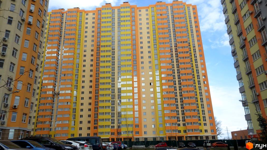 Яскравий житловий комплекс: висотні будинки з яскравим фасадом в помаранчево-жовтих тонах. На прибудинковій території видно припарковані автомобілі.