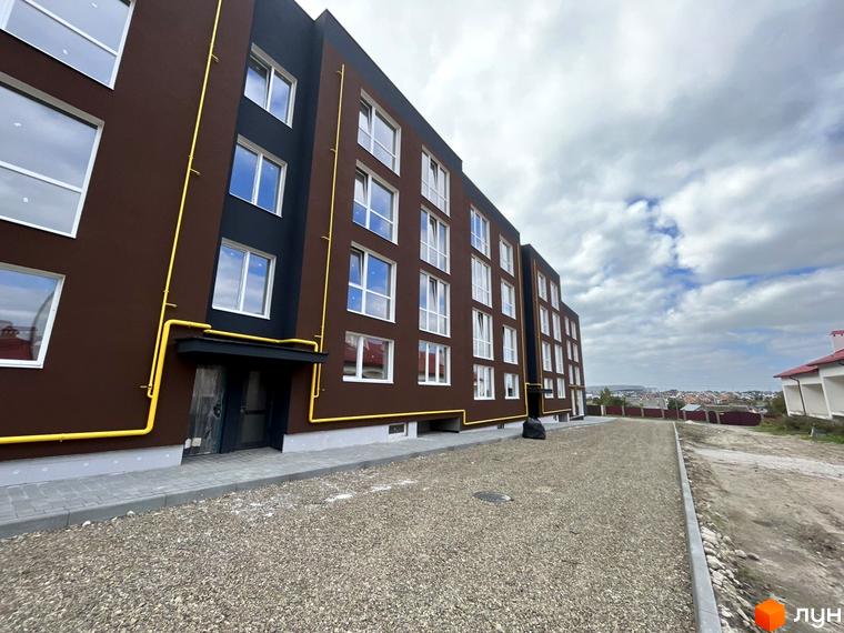 Зовнішній вигляд будинку ЖК Зубра Дім з коричневими стінами та великими вікнами. Поряд з будинком розташована прибудинкова територія з гравієм та тротуаром.