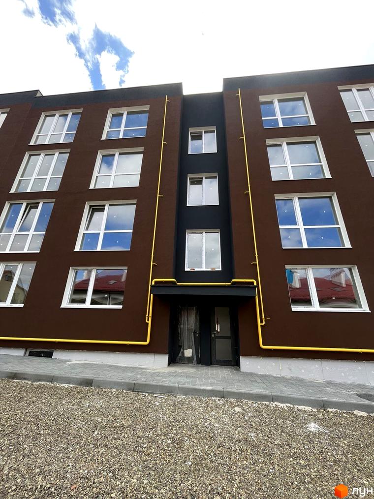 Зовнішній вигляд будинку Житловий комплекс Зубра Дім з коричневим фасадом, білими вікнами та жовтими трубами. На передньому плані - бруківка та гравій.