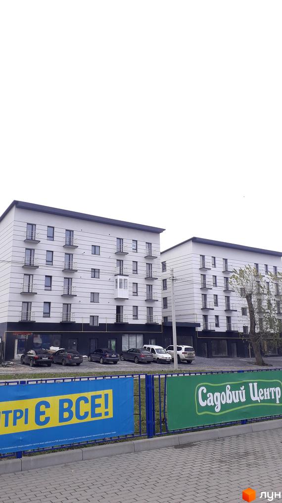 Два багатоповерхові будинки з балконами та світлими фасадами. Перед ними парковка та огорожа з рекламними банерами.