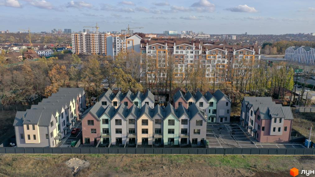 Таунхауси New Smart 11 - це новий будівельний проєкт з різнокольоровими фасадами та сірими дахами, розташований на тлі багатоповерхових будинків та міського пейзажу.