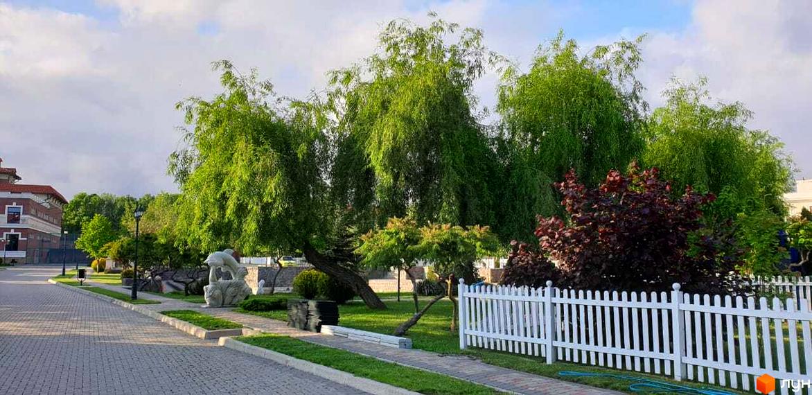 Зовнішній вигляд Котеджного містечка Аріадна з великими зеленими деревами та білим парканом. На території розташовані скульптури та доглянуті газони.