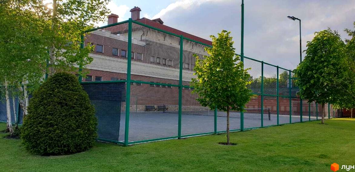 Зовнішній вигляд Котеджного містечка Аріадна з тенісним кортом. На передньому плані зелений газон, дерева та кущі.