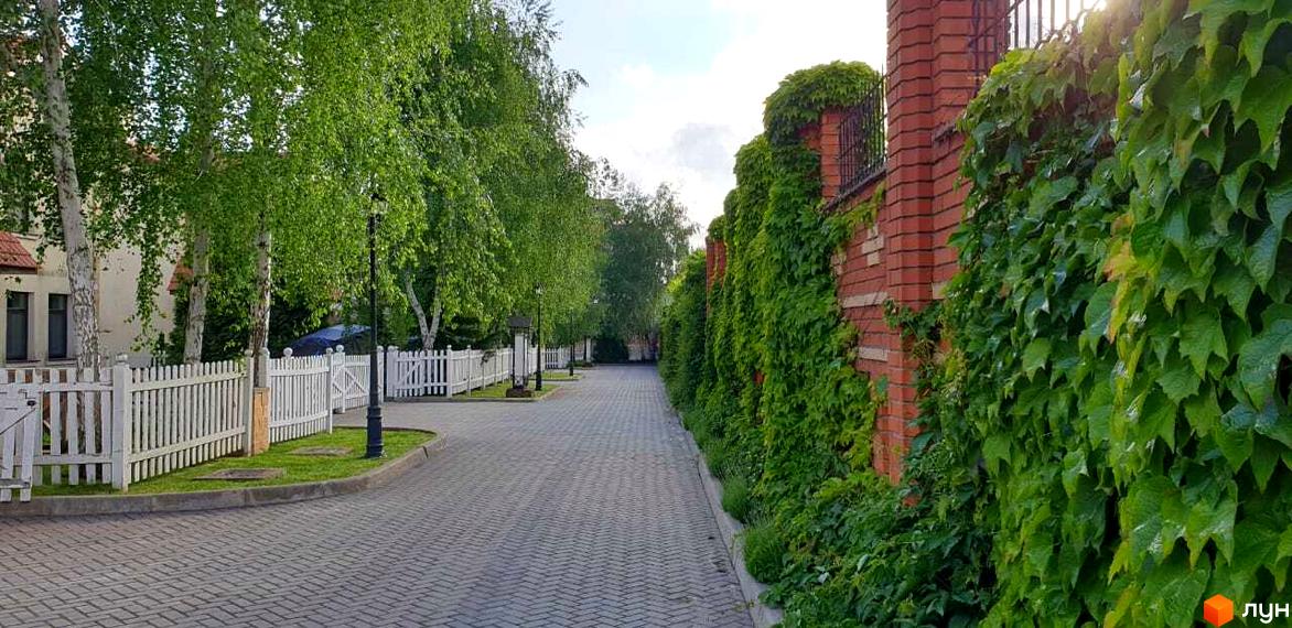 Вигляд вулиці в Котеджному містечку Аріадна з бруківкою та зеленими насадженнями. Цегляна огорожа, частково вкрита рослинністю, та білий паркан вздовж дороги.