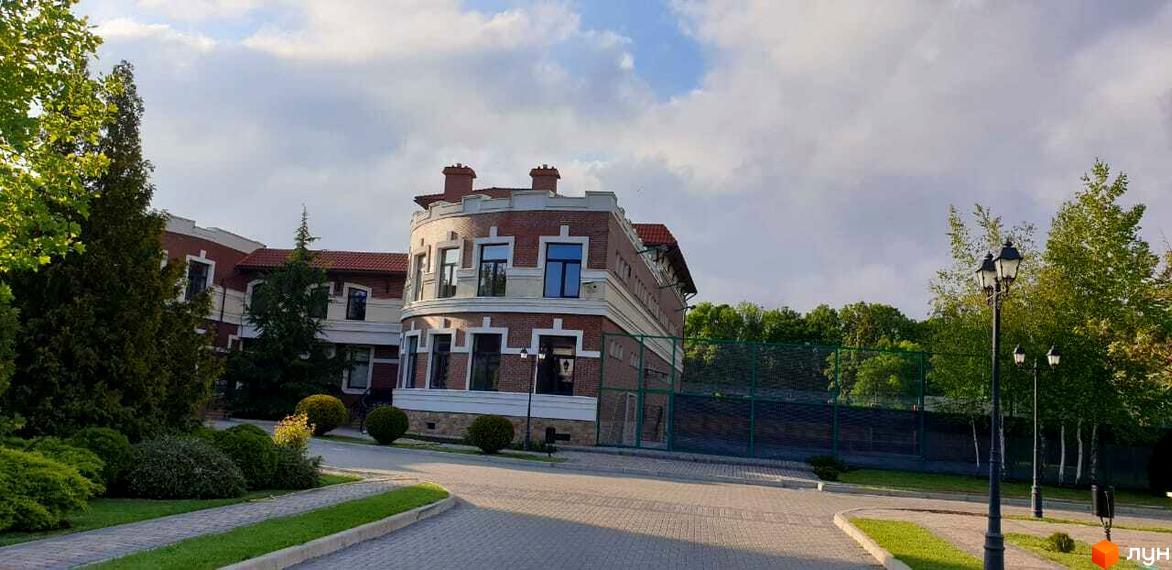 Зображення Котеджного містечка Аріадна: триповерховий будинок з червоної цегли з білими декоративними елементами. Навколо зелені насадження та доріжки.