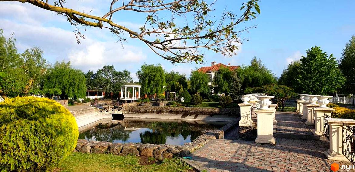 Зображення Котеджного містечка Ariadna з озером та ландшафтним дизайном. На задньому плані будинок з червоним дахом та зелені насадження.