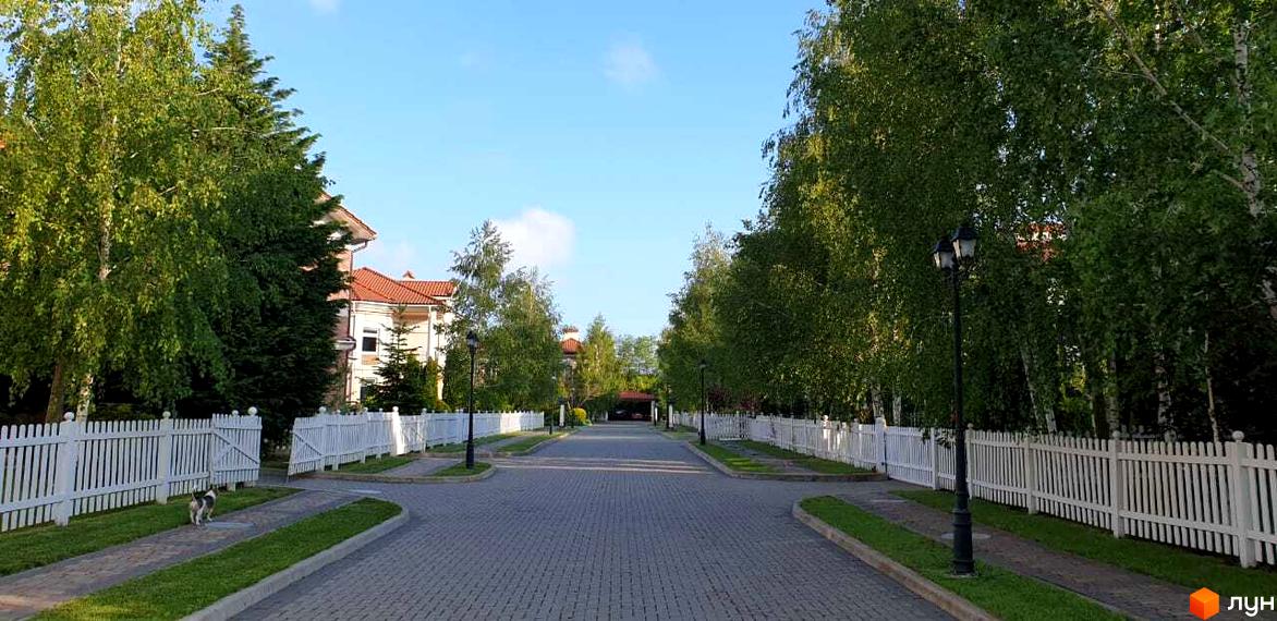 Зображення Котеджного містечка Аріадна з білими парканами та деревами вздовж дороги. Вигляд на житлові будинки з червоними дахами та зелену прибудинкову територію.