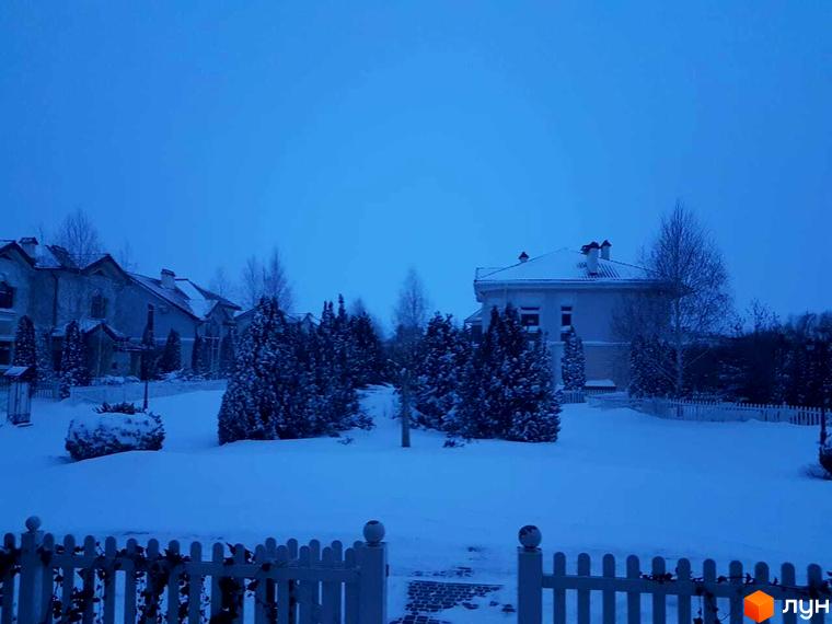 Зимовий пейзаж: будинки Котеджного містечка Аріадна під снігом. На передньому плані засніжена територія з ялинками та декоративною огорожею.