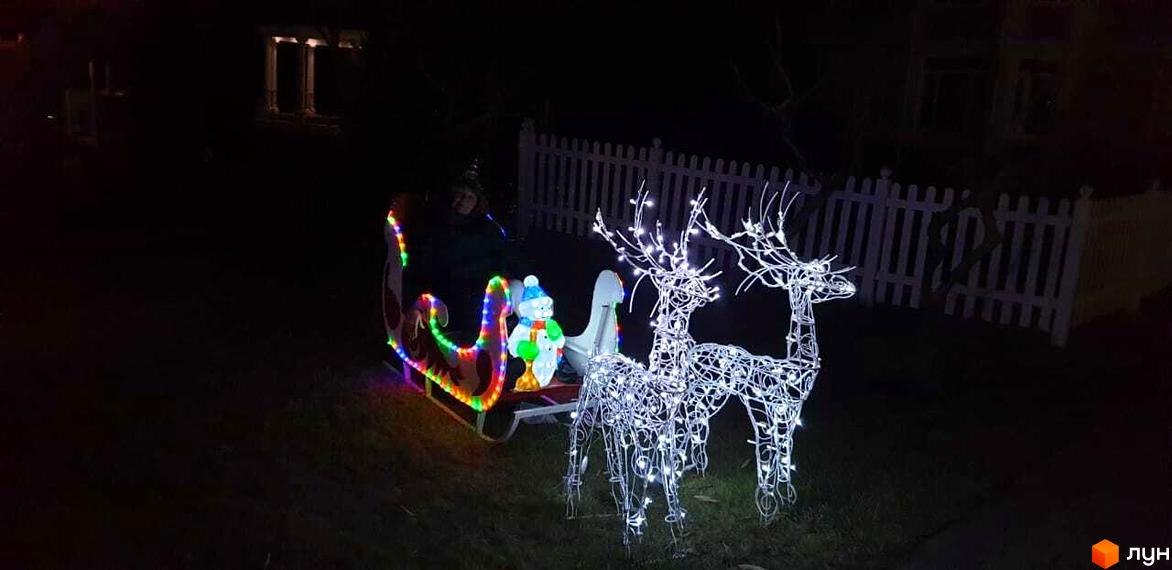 Новорічна ілюмінація: сани з фігурою сніговика та двома оленями на фоні білого паркану. Святкова атмосфера біля Котеджного містечка Аріадна.