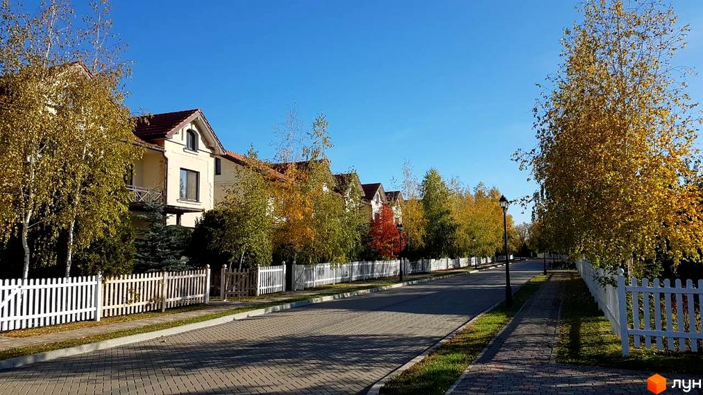 Котеджне містечко Аріадна: фасади будинків з терасами та червоними дахами, вздовж вулиці ростуть дерева з жовтим листям, білі паркани.