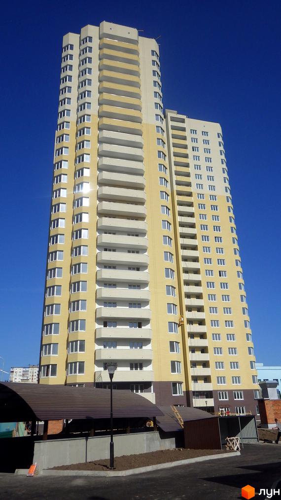 Високоповерховий житловий комплекс Гранд жовтого кольору з білими балконами на фоні блакитного неба. Поруч розташована прибудинкова територія з навісами та ліхтарем.