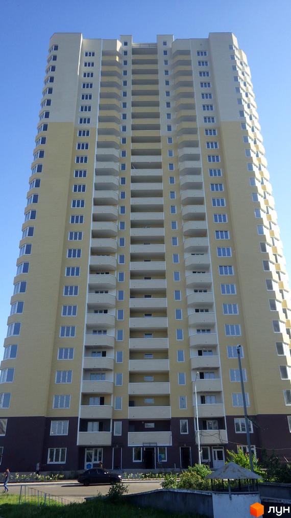 Високоповерховий житловий комплекс Гранд з жовто-білим фасадом та численними балконами. Навколо будинку зелена зона та проїжджа частина з автомобілями.