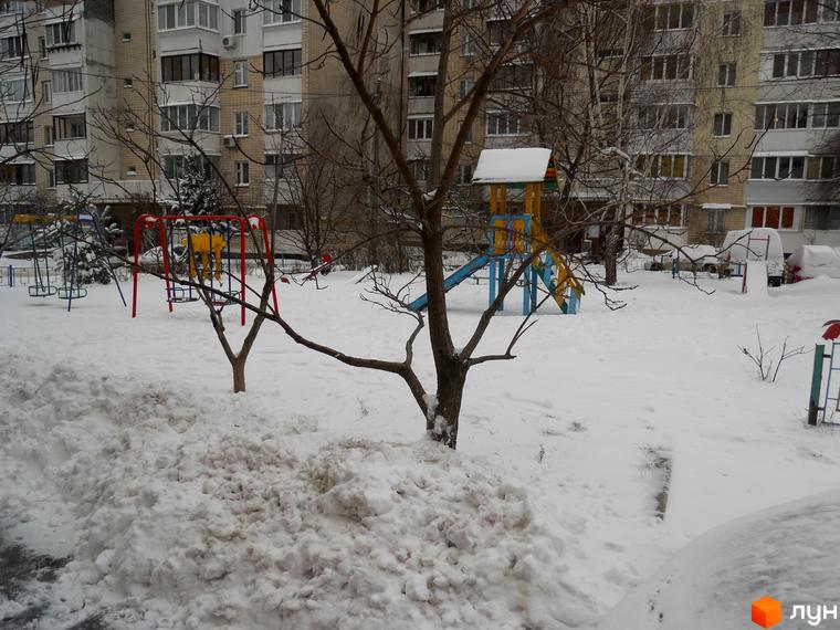 Зимовий пейзаж Житлового комплексу Мінський: сніг вкриває дитячий майданчик та дерева біля багатоповерхового будинку.
