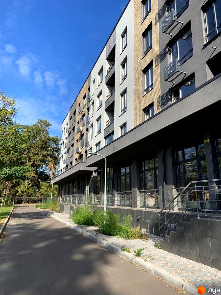 Багатоповерховий Апарт-комплекс в Пущі Водиці з фасадом у світлих, бежевих та темних тонах. Прилегла територія має доріжку, зелені зони та дерева.