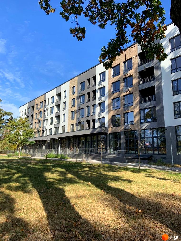 Багатоповерховий Апарт-комплекс в Пущі Водиці з фасадом з комбінованих матеріалів та великими вікнами. Перед будинком зелений газон та дерева, що створюють тінь.