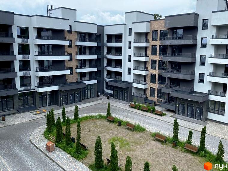 Апарт-комплекс в Пущі Водиці: багатоповерхова будівля з сірими фасадами та балконами. Прибудинкова територія має бруківку, дерева та лавки.