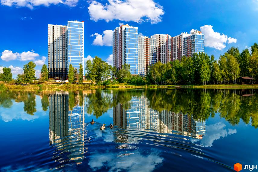 Житловий комплекс Причал 8 з високими будинками та блакитними вікнами, що відбиваються у воді озера. Навколо зелені дерева та хмарне небо.