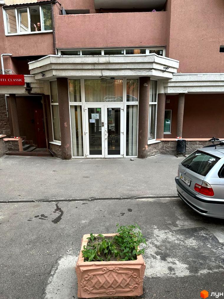 Вхід до житлового комплексу Елегант з сучасним фасадом та прибудинковою територією, оформленою рослинами.