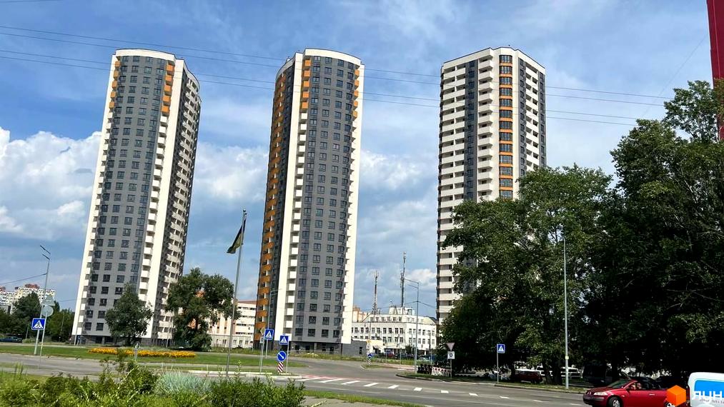 Житловий комплекс Райдужний – це три високі багатоповерхові будинки сірого та білого кольору з помаранчевими елементами. Навколо новобудов зелена зона та дорога.