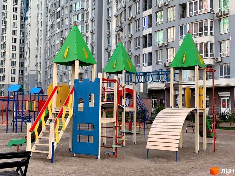 Дитячий майданчик перед багатоповерховим будинком ЖК Каховська. На майданчику різнокольорові гірки, гойдалки та пісочниця. Будинок має сірий фасад та багато вікон.